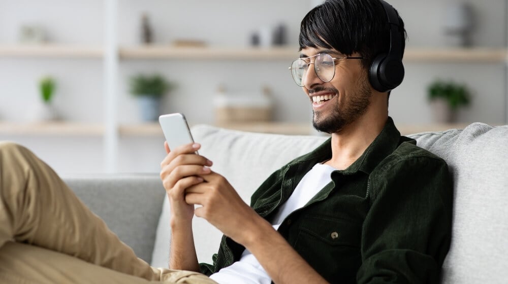 man wearing headphones sitting on grey couch holding phone and smiling